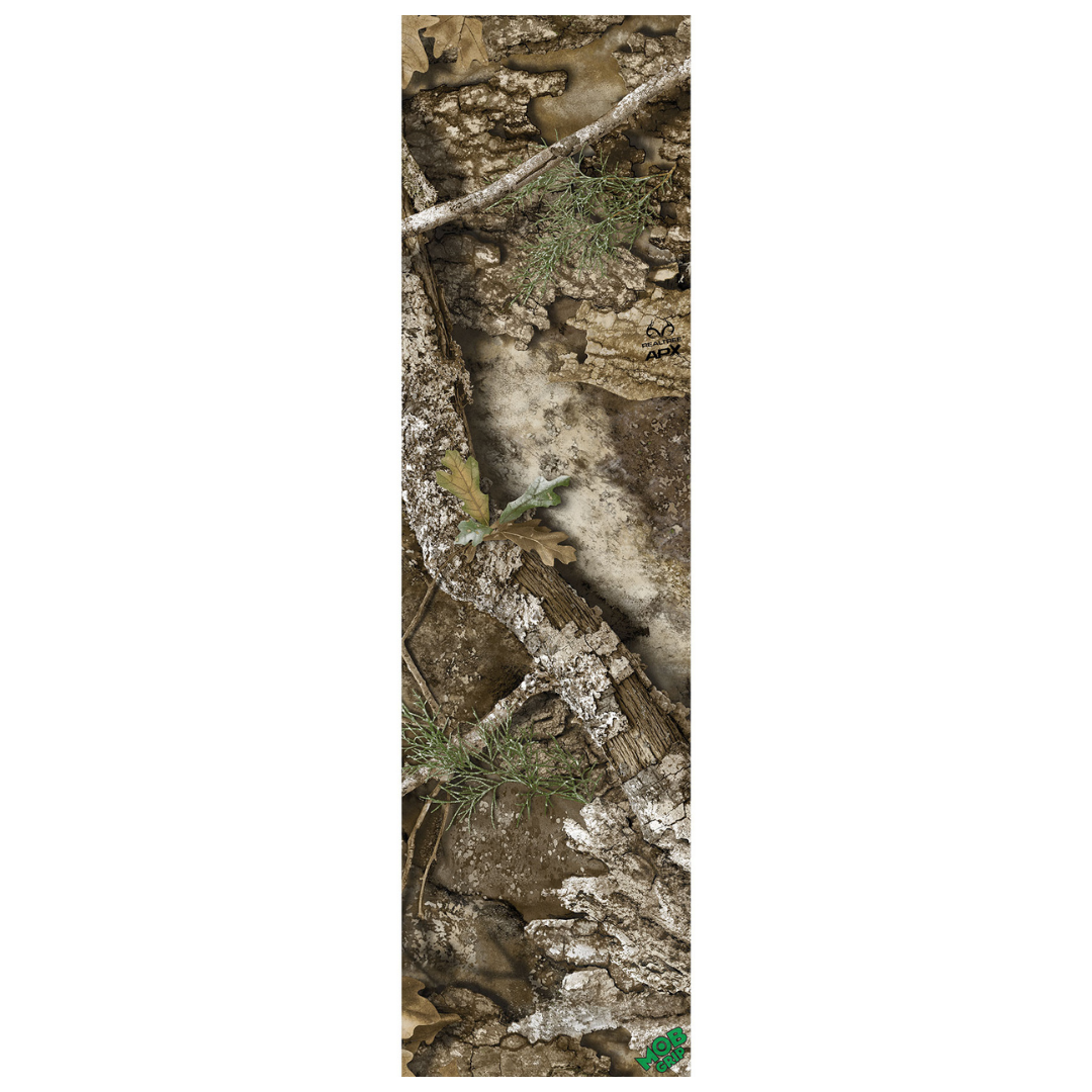 A narrow rectangular camouflage-patterned item, likely a sleeve or cover, featuring a realistic woodland print with bark, branches, leaves, and dirt in muted brown, gray, beige, and green tones. A small green logo appears at the bottom right.