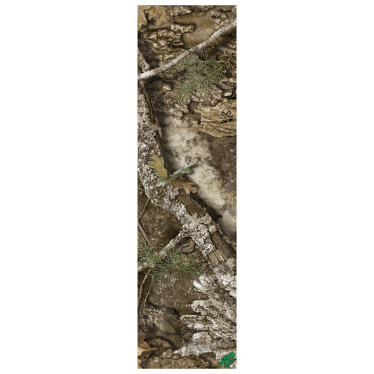 A narrow rectangular camouflage-patterned item, likely a sleeve or cover, featuring a realistic woodland print with bark, branches, leaves, and dirt in muted brown, gray, beige, and green tones. A small green logo appears at the bottom right.