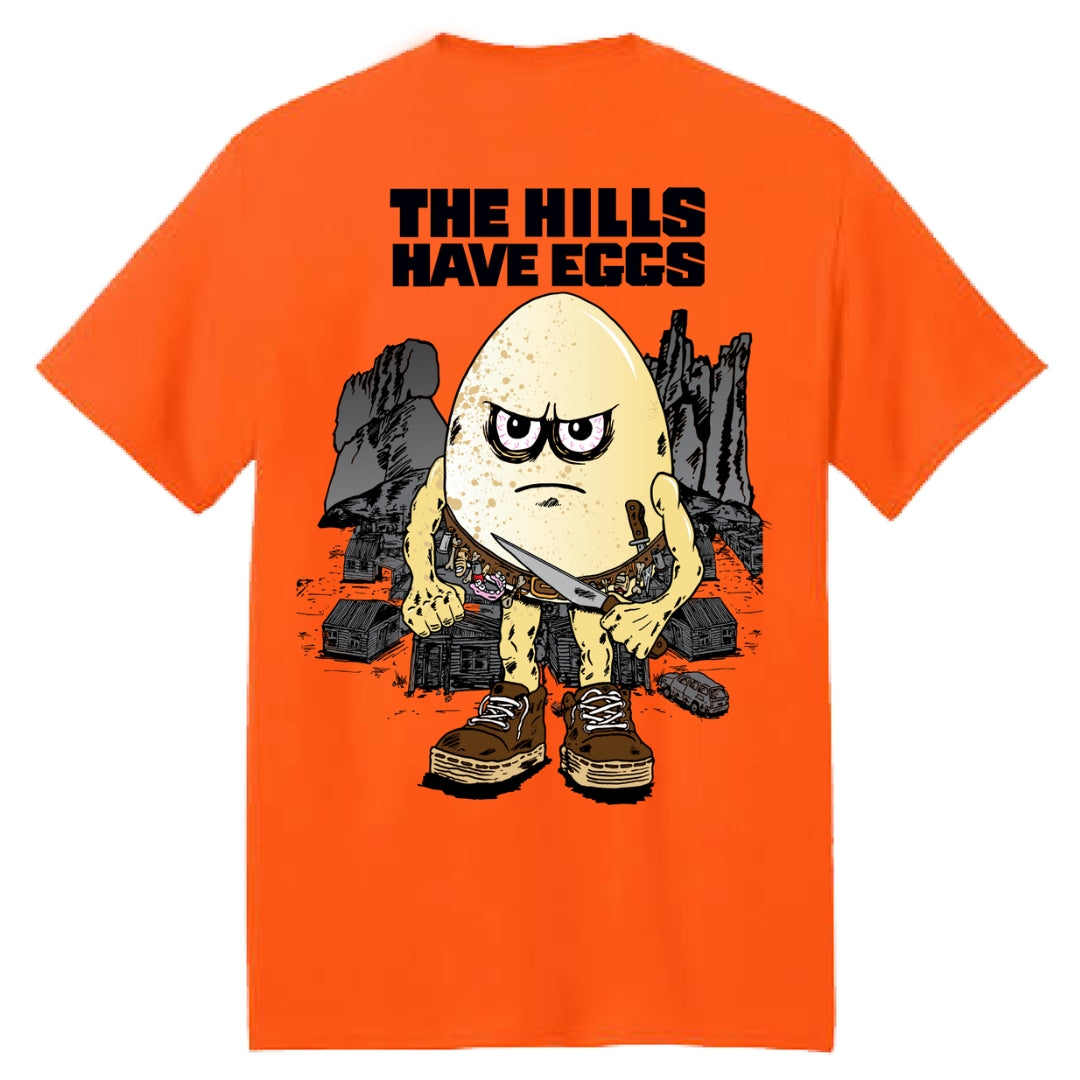 Orange t-shirt with a graphic design of an egg character holding a knife and text 'THE HILLS HAVE EGGS' on the back.