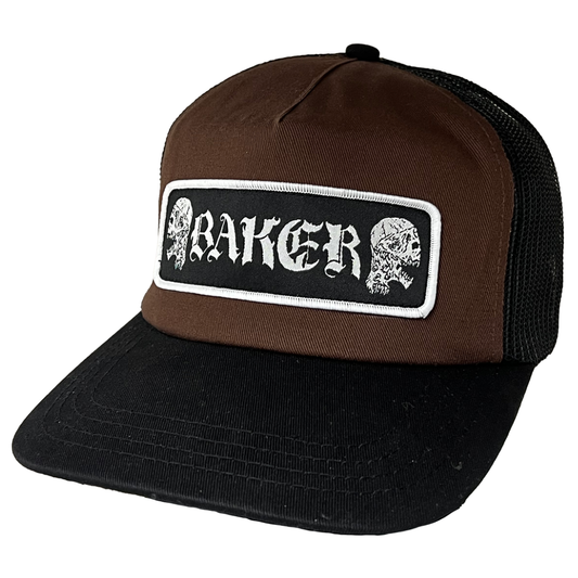 A brown and black trucker hat with a rectangular front patch featuring the word 'BAKER' in white gothic lettering between two white skulls, shown on a plain white background.