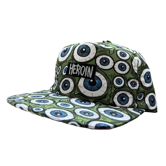 A flat-brim baseball cap featuring a green and white 'gory eyes' pattern, with a black embroidered patch on the front that reads 'C HERoin'.